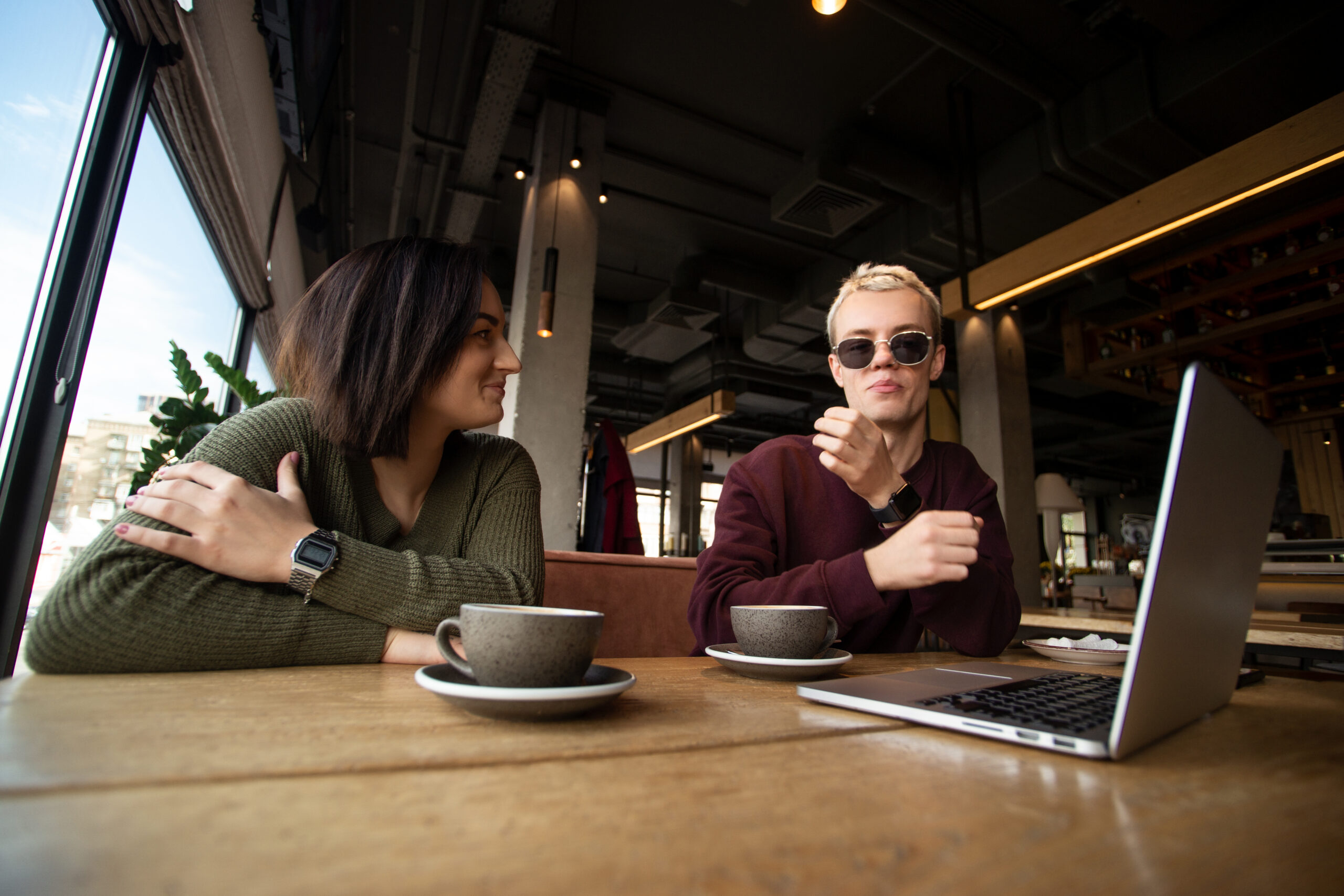 Couple working together in cafe. Young brunette woman smiling and looking at cool blonde man in black round glasses. Gray laptop on table. Two cups of coffee. Freelance working concept. Couple working together in cafe. Young brunette woman smiling and looking at cool blonde man in black round glasses. Gray laptop on table. Two cups of coffee. Freelance working concept