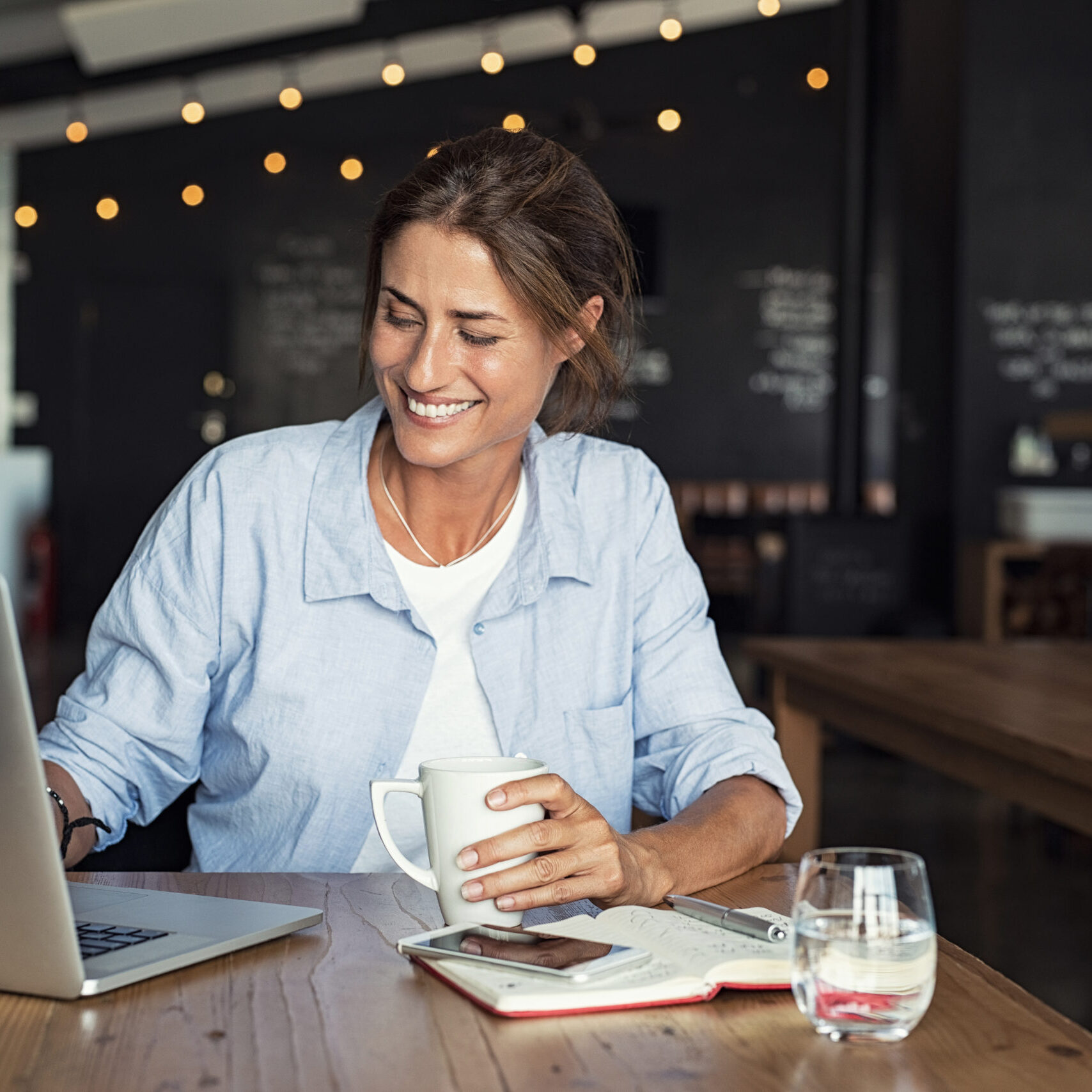 Mature woman working on laptop Smiling woman sitting in cafeteria holding coffee mug and working on laptop. Businesswoman checking email on laptop. Beautiful middle aged woman smiling and using laptop at cafe while drinking a cup of tea.